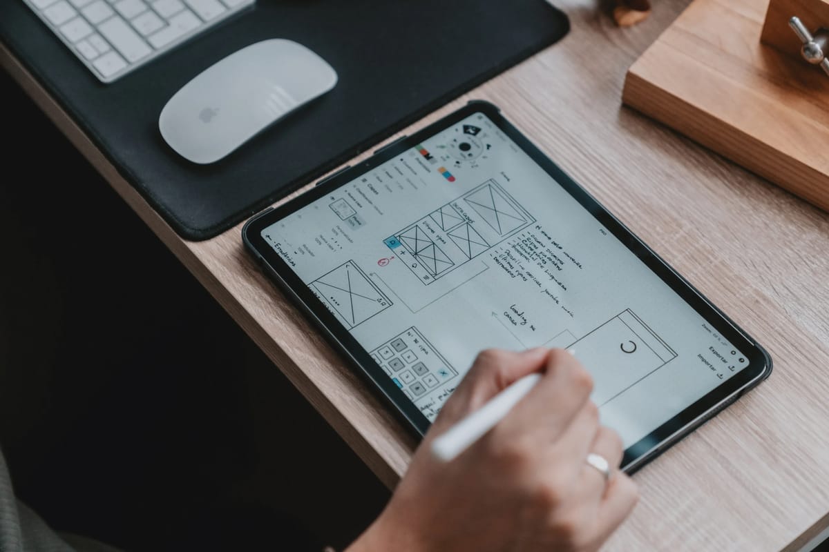 En hånd som bruker Apple Pencil på en iPad som ligger på et trefarget skrivebord. På skjermen vises wireframes og UI-skisser for en nettside eller app. Ved siden av iPaden ses en hvit Apple Magic Mouse og tastatur på en svart musematte.